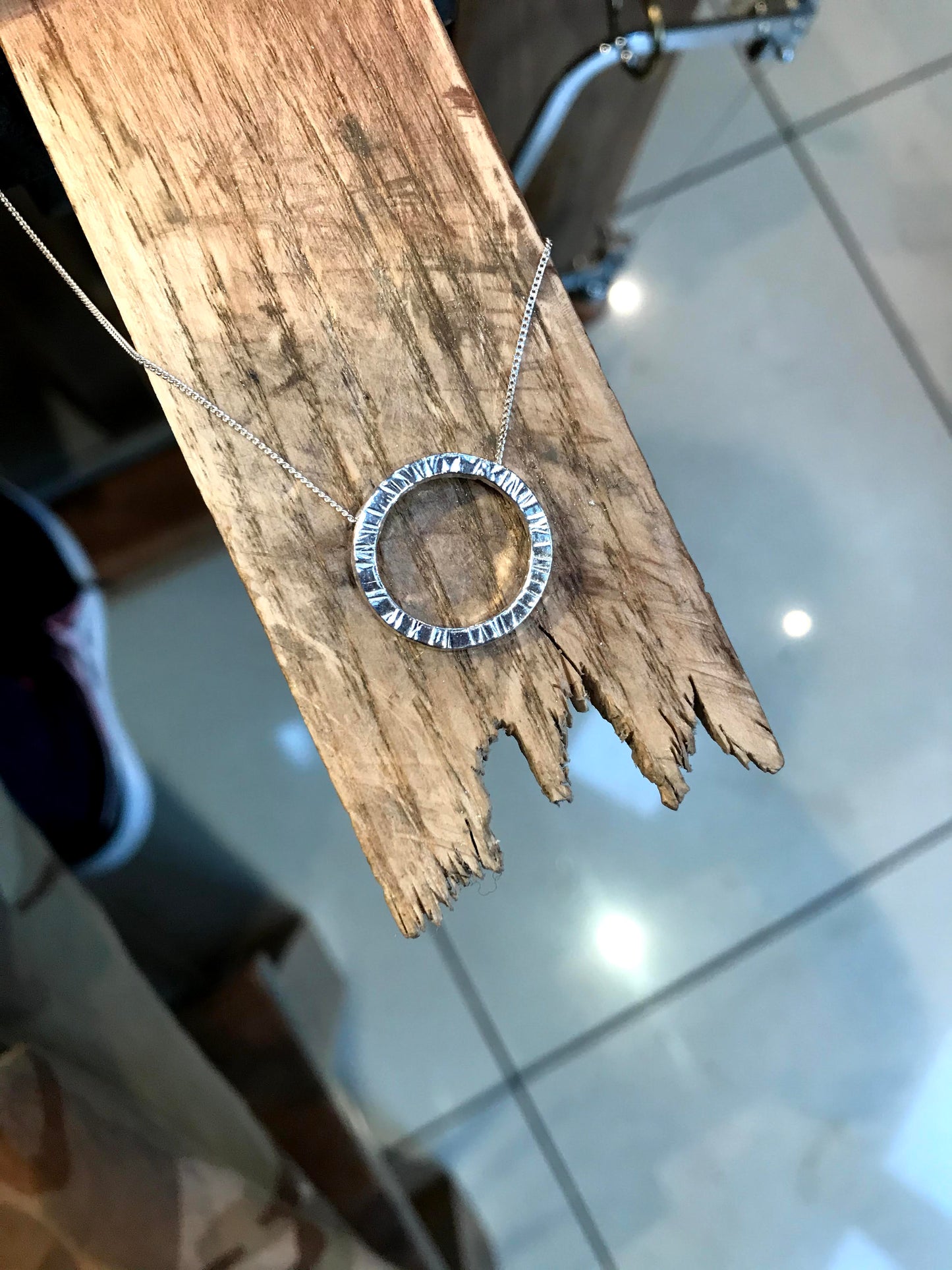 Handmade sterling silver 20cm circle of silver made from 2mm square wire suspended on a hidden bale behind the circle. The circle appears to float on a sterling silver diamond cut curb chain. Traditional wooden bench peg