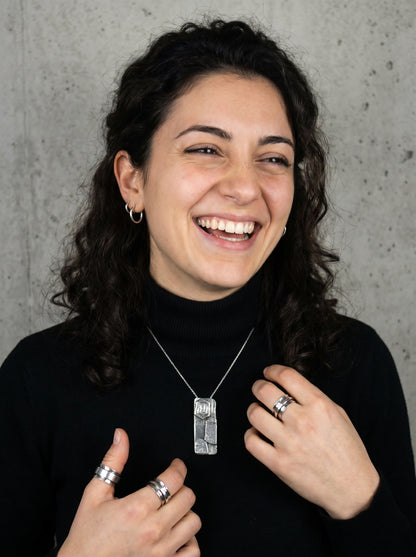 Woman wearing a necklace and rings, smiling against a concrete wall.