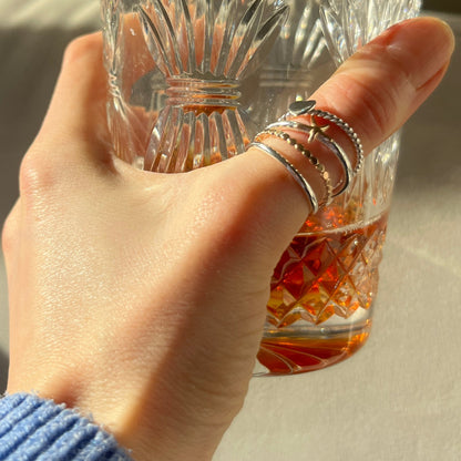 Hand holding a glass of amber liquid with a textured glass, wearing multiple silver rings on a neutral background.