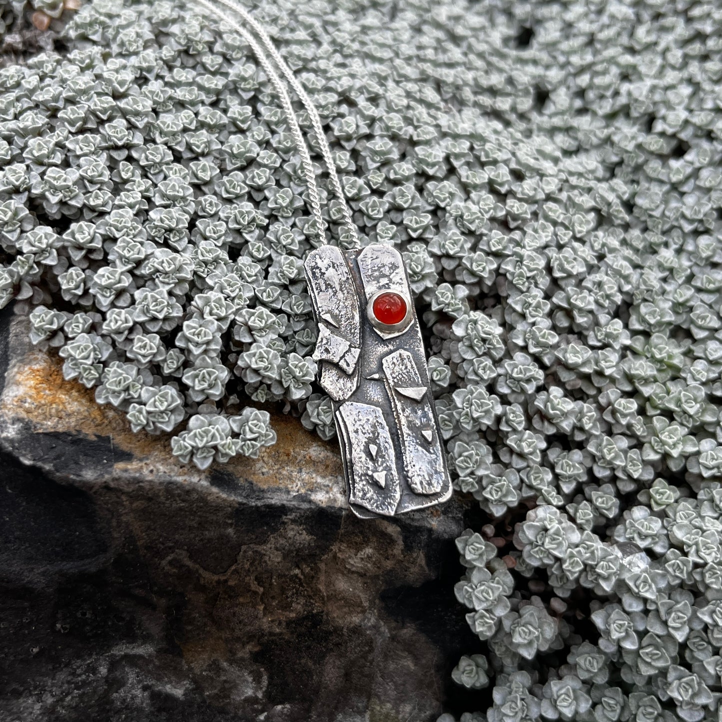 Silver pendant with a red gemstone on a rocky surface