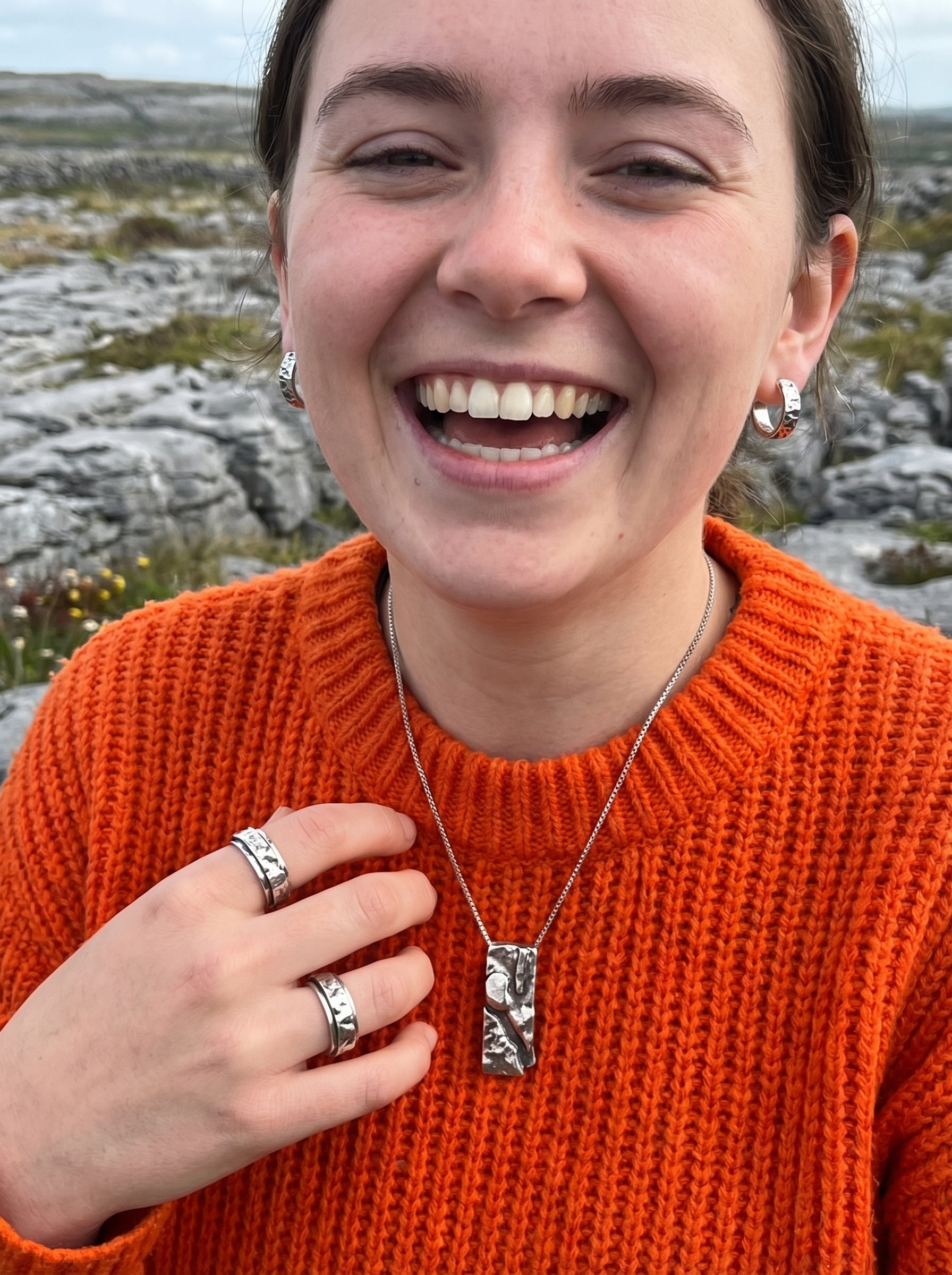 Person wearing an orange sweater with jewelry, standing in a rocky landscape.