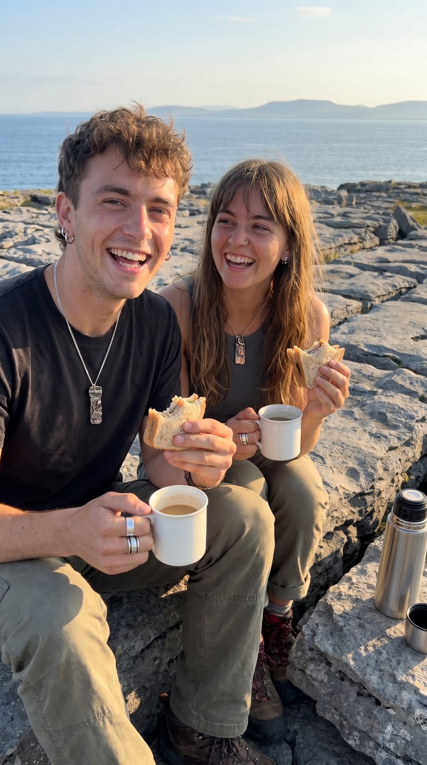 Two people sitting on rocks by the ocean, enjoying coffee and a sandwich.