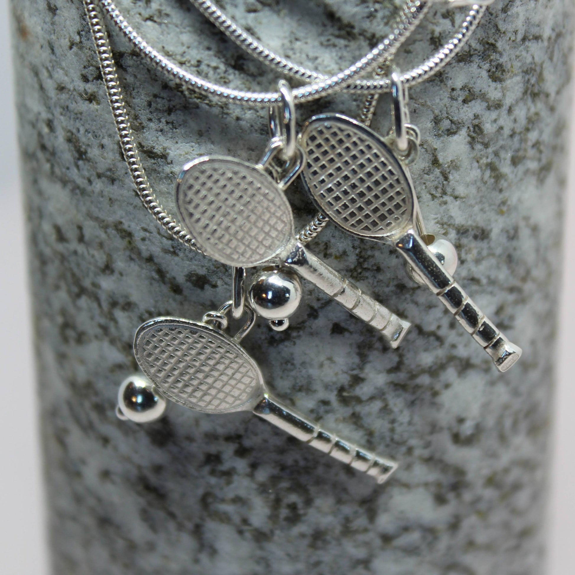 Three sterling silver tennis racket pendants on a grey granite rock